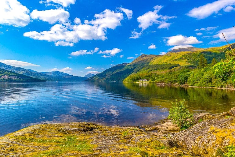 blue lake with green rolling hills beside and puffy clouds in blue sky above in loch lomond scotland.