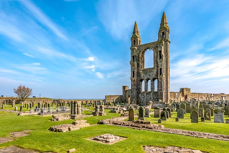 church stone ruins with grave stones in green grass cemetery with blue sky behind.