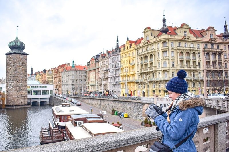 Frau mit Wintermütze steht auf Brücke in Prag mit bunten Gebäuden im Hintergrund