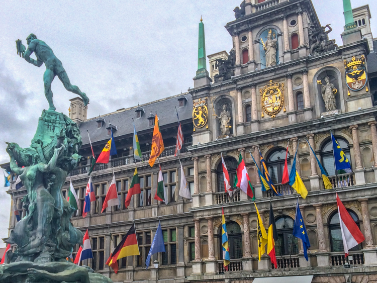 colourful flags on large old building in antwerp belgium with statue in front.