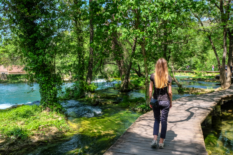 girl walking through trees on wooden path with river underneath krka national park