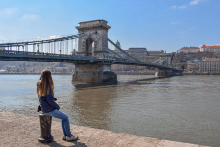 girl sitting on river edge with bridge behind her on eastern europe itinerary