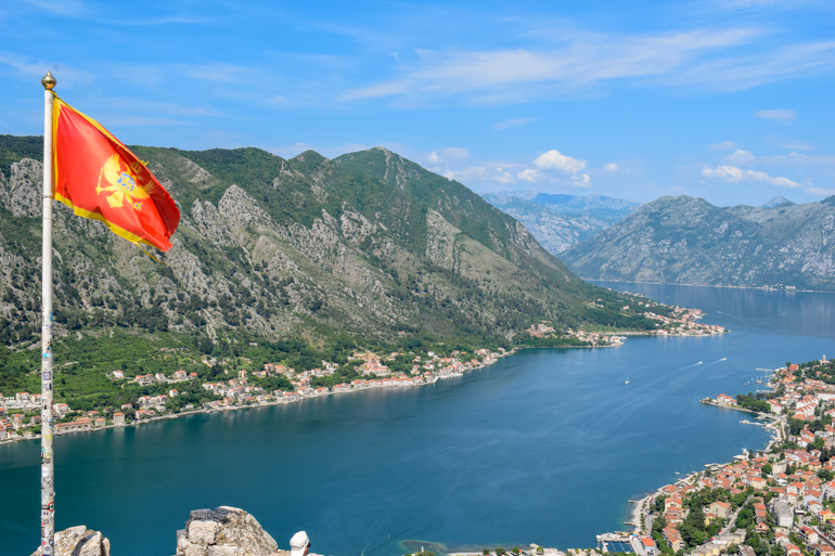 montenegro flag flying with blue bay of kotor and old town below in background.