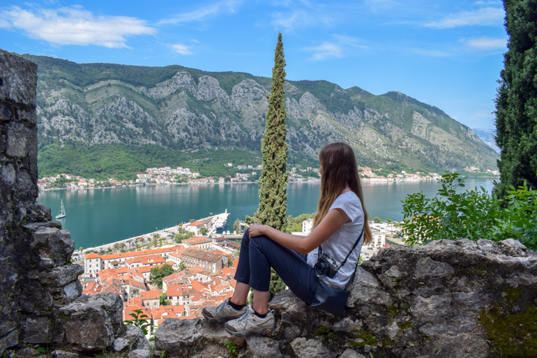 girl in white shirt sitting on fortress wall with bay of kotor in background.
