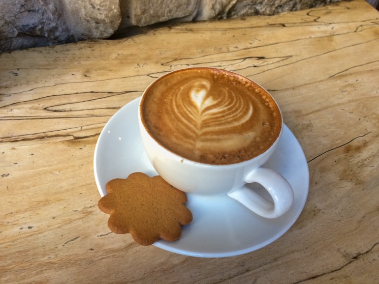 latte in white mug and cookie on wooden table with rock wall coffee shops in edinburgh