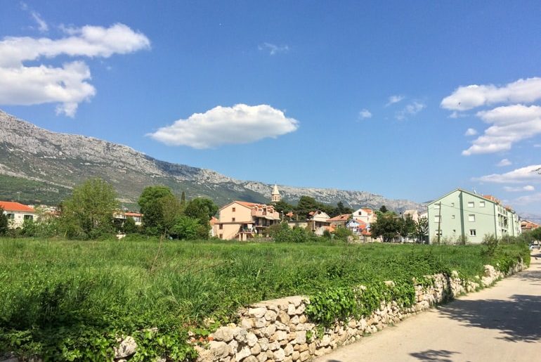 road with green field and old buildings beside with mountain in background in croatia road trip