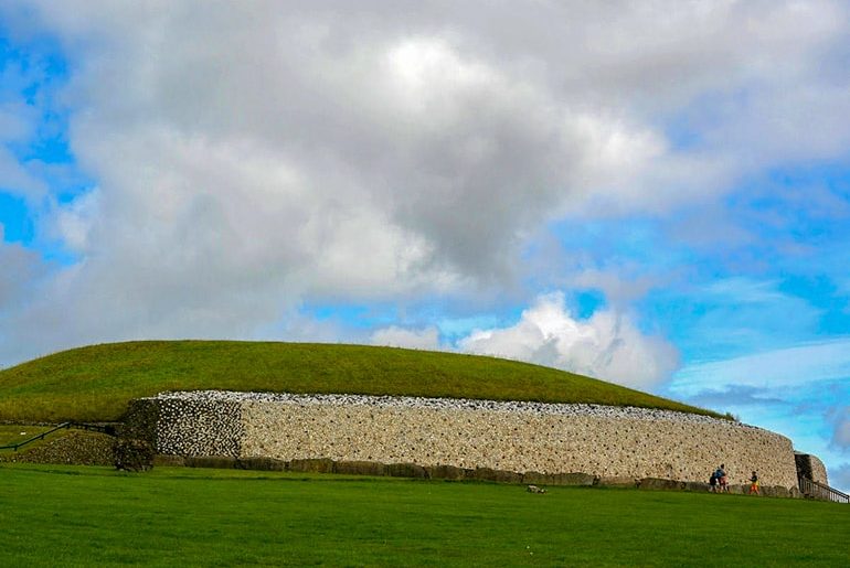 newgrange round stone and green grass monument with cloudy blue sky ireland travel tips