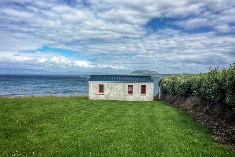 small cottage with green lawn and crashing ocean behind with blue sky ireland travel tips