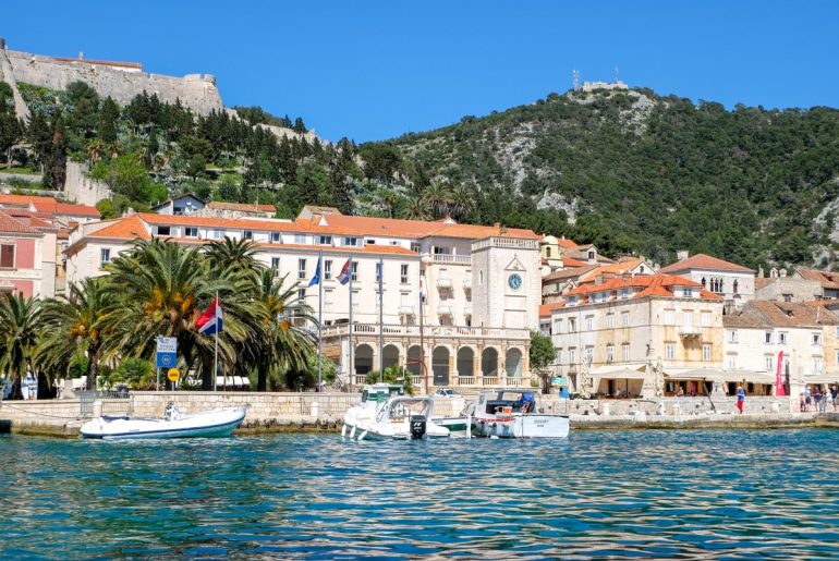 white building with clock and parked boats in front of blue sea and harbour in hvar