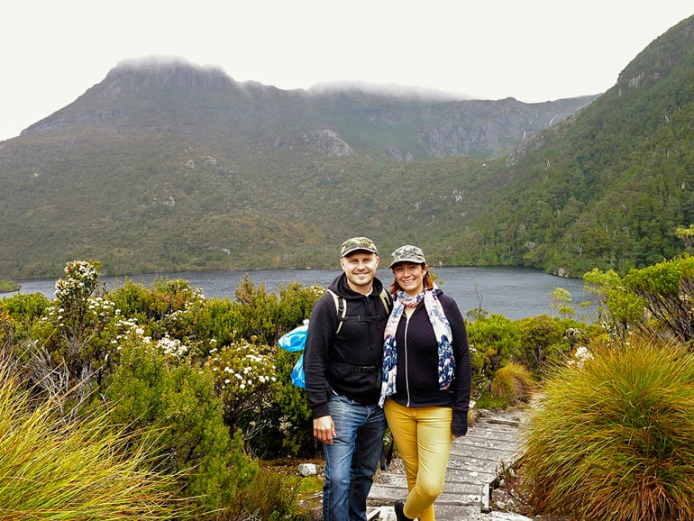 Two people on wood walkway with green shrubs and mountains in background