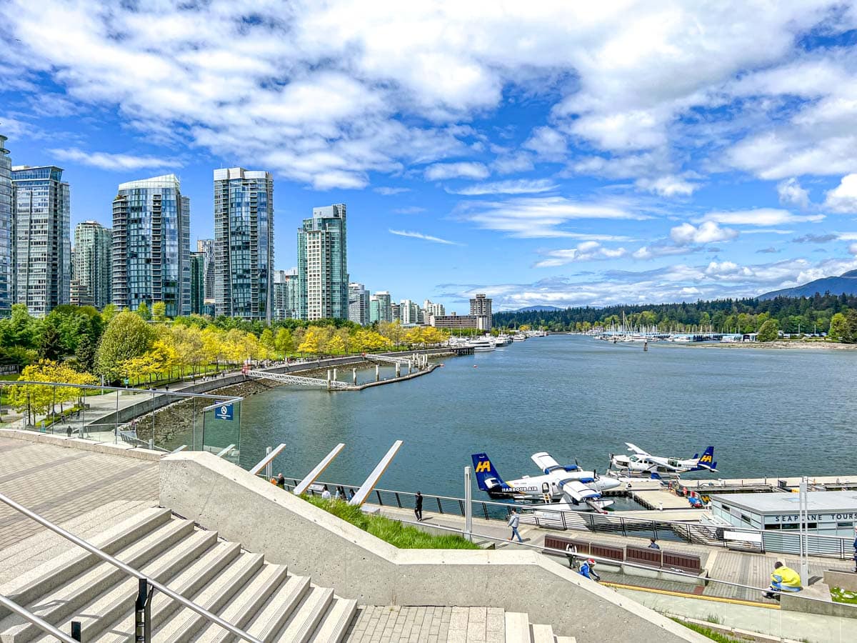 tall glass and steel buildings along water edge in vancouver with trees and mountains in the distance behind.