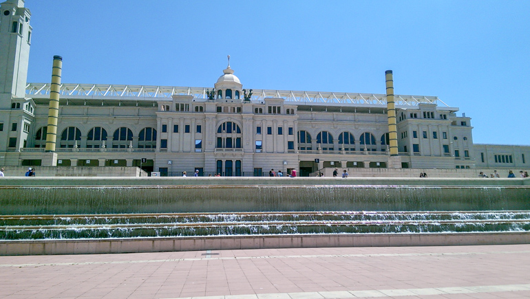 long stadium building with fountain in front and blue sky above
