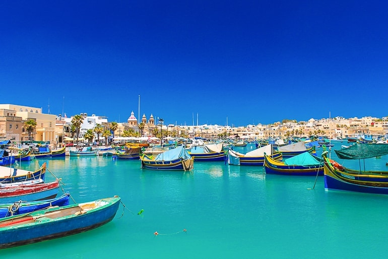 covered fishing boats on blue water with fishing village in Malta behind on shore