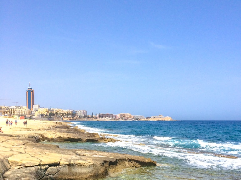 rocky shoreline with blue ocean and buildings in background in malta