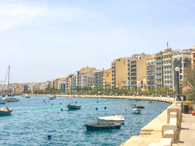 Uferpromenade mit Bäumen entlang des blauen Hafens mit Booten und Häusern im Hintergrund in Sliema, Malta