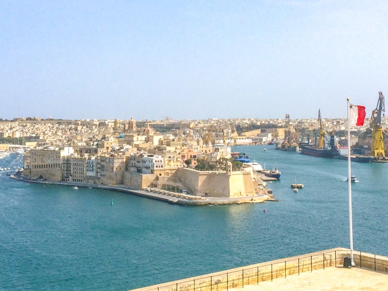 pier with old buildings in blue harbour with flag pole in front in valletta malta