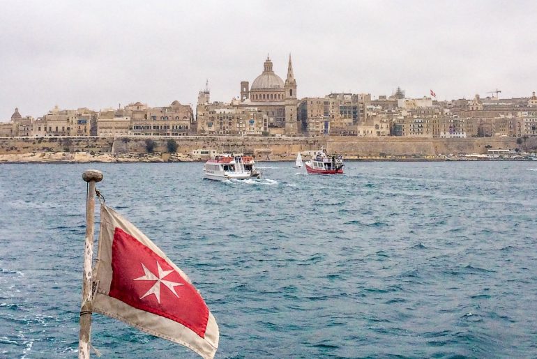 maltese flag with boats in water and city in background travelling to malta