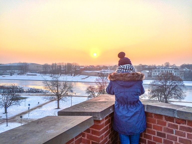 girl in winter hat and jacket seen from behind watching orange sunset.