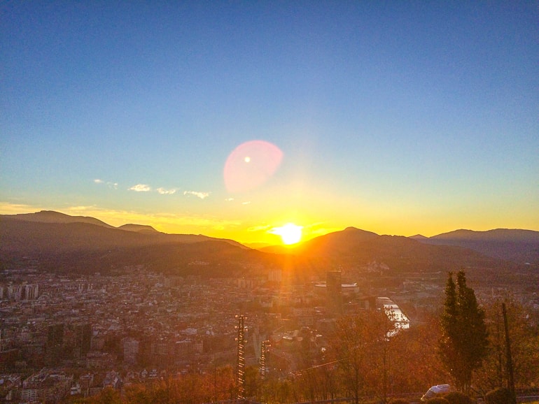 sun seen above mountain range setting over city with river in middle.