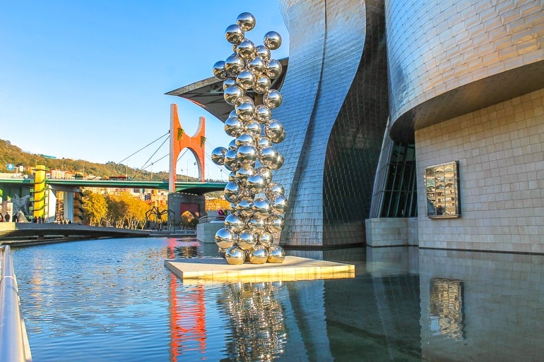 steel balls in sculpture over water fountain with museum building behind.