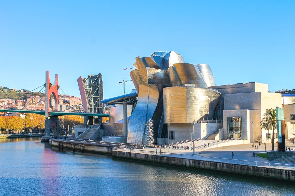 metallic museum building with river flowing past and red bridge behind.