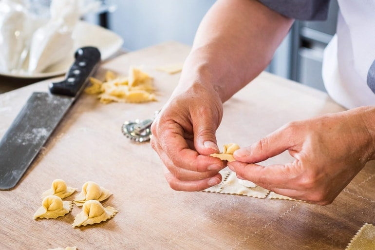 hands shaping pasta on wooden counter with knife