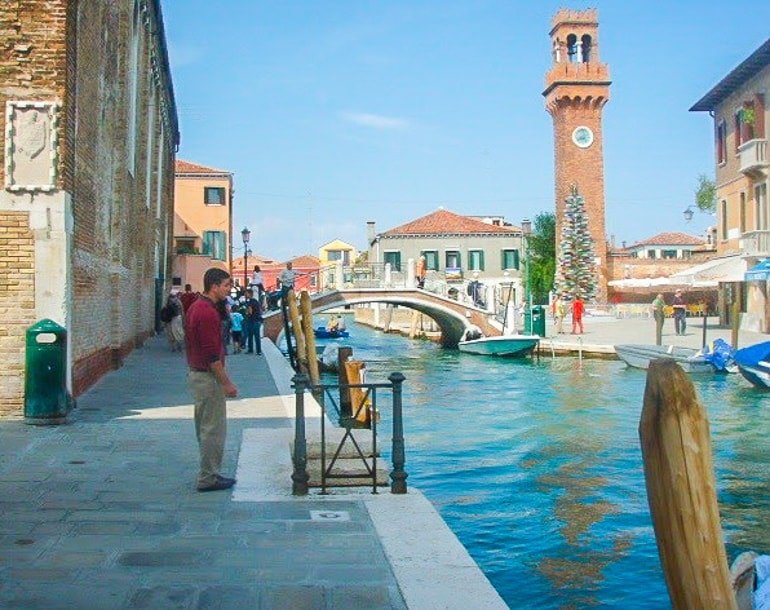 man standing beside water on promenade with tower behind murano