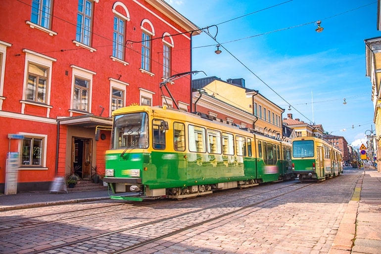 green street cars pass by red building with cobblestone street underneath in helsinki.