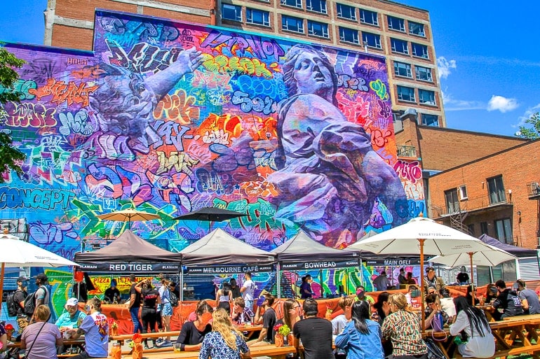large wall with colourful mural and people sitting at tables with umbrellas in front.