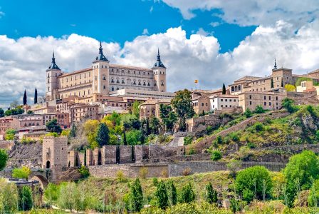 square castle with spires on hillside with green trees below beautiful cities in spain toledo