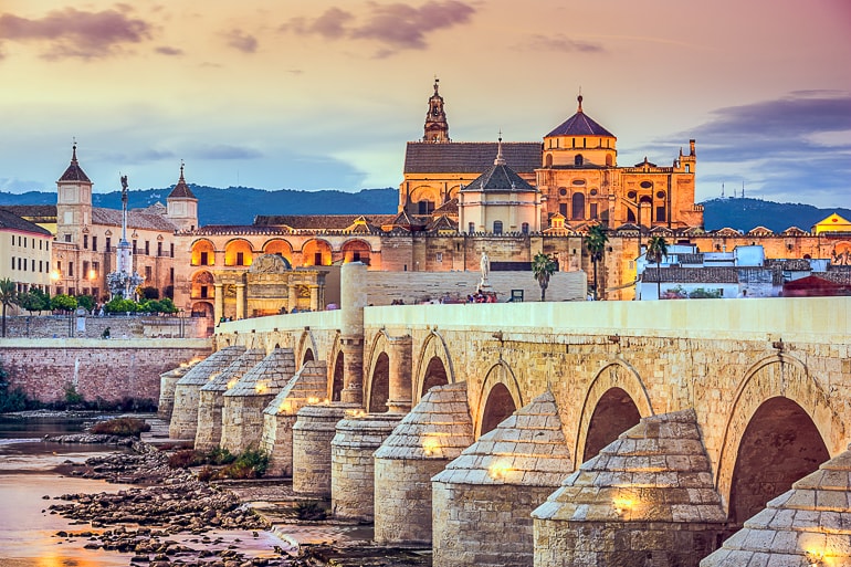 Steinbrücke im Vordergrund mit orange beleuchteter Kirche auf Hügel dahinter Cordoba Spanien
