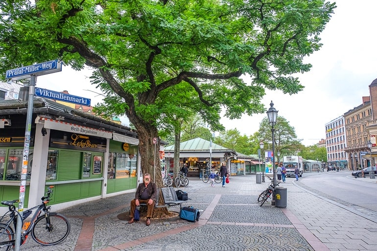 Mann sitzt neben Baum mit geschlossenen Marktständen im Hintergrund München Viktualienmarkt
