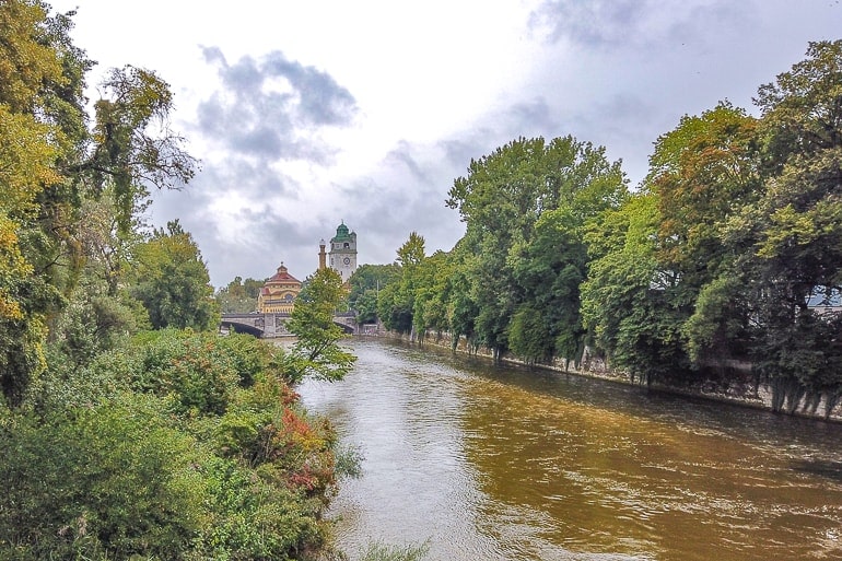 green trees lining brown river in munich with buildings behind.