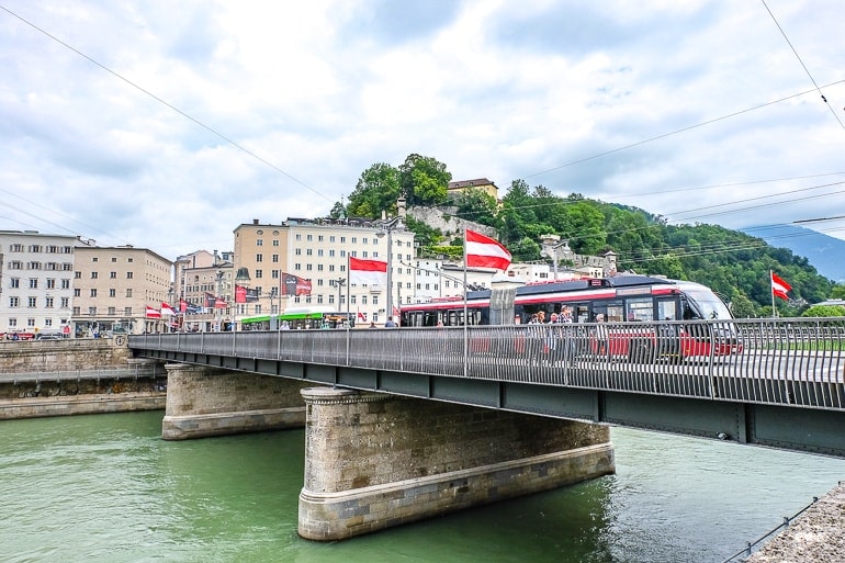 Rote Straßenbahn überquert Brücke in Salzburg mit Gebäuden im Hintergrund.