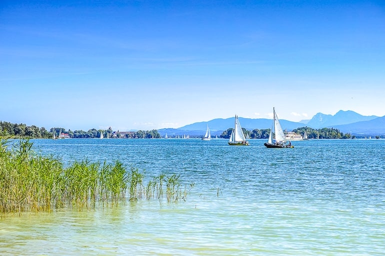 blue lake with green grass beside and sailboats in distance chiemsee