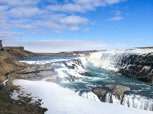 Blauer Wasserfall mit Gischt und schneebedeckte Felsen im Vordergrund bei blauem Himmel