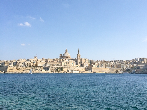 view of sandstone buildings of old town in distance from across blue water in Malta.