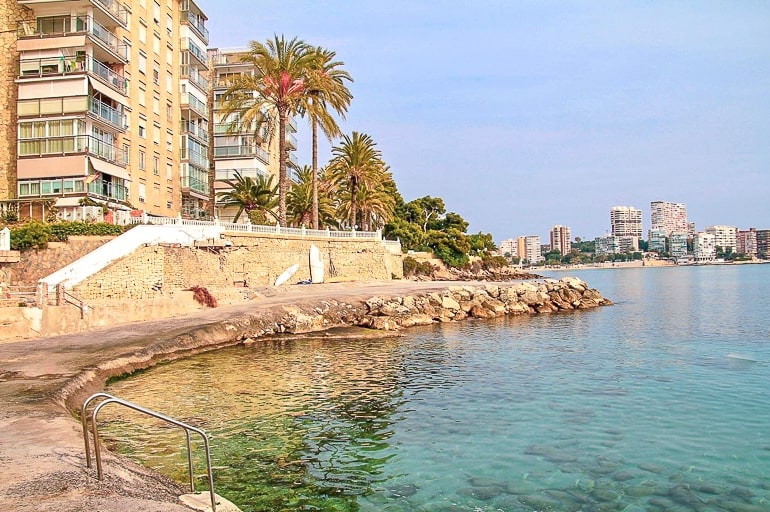 pathway along blue coast with trees and buildings behind in alicante