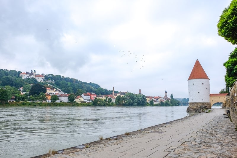 river pathway with tower and green shoreline with houses behind in distance in passau innstadt