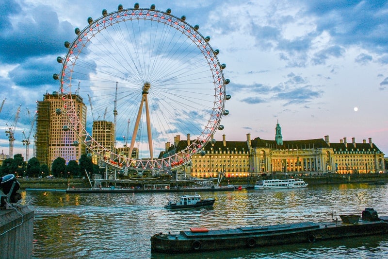 large ferris wheel standing with river thames in front with boats cruising past at dusk.