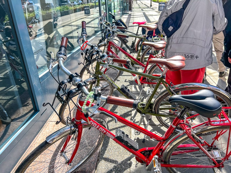 red and green bikes lined up on sidewalk with glass building and man in jacket behind.