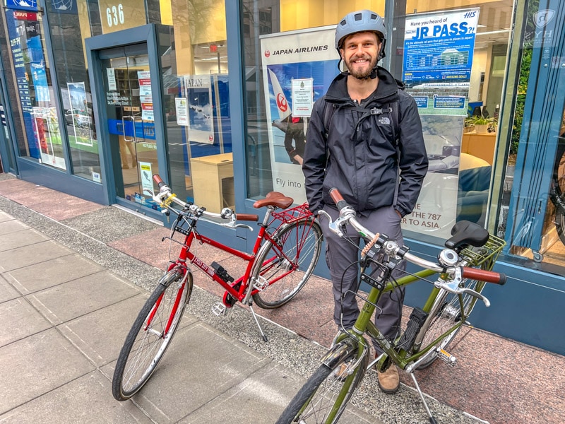 Ein Mann mit Helm und Jacke steht zwischen einem roten und einem grünen Fahrrad auf dem Bürgersteig.