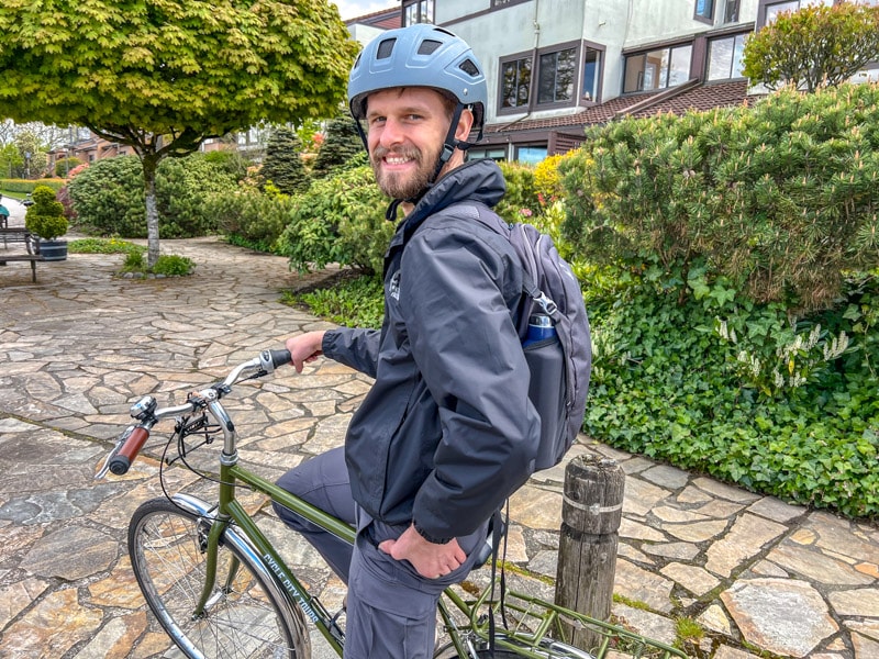Mann mit Bart sitzt auf dem Fahrrad mit Helm und grünen Büschen im Hintergrund.