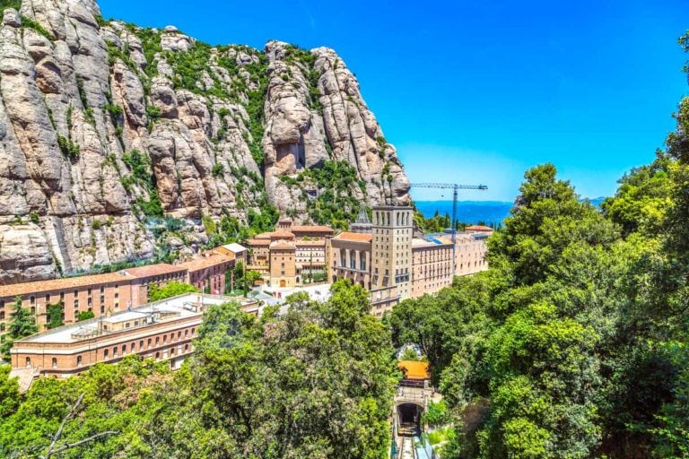monastery of montserrat nestled in base of mountain cliff with green trees in foreground.