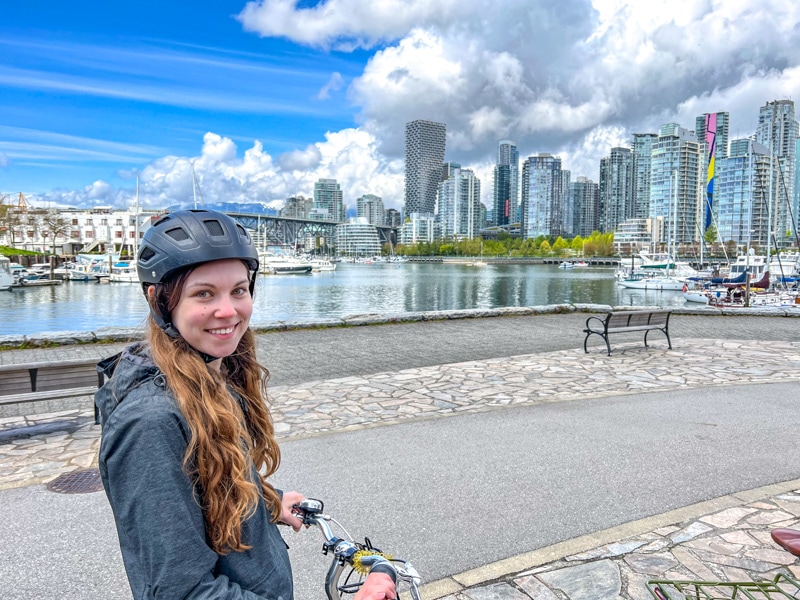 Eine Frau mit Helm steht neben einem Fahrrad mit den Gebäuden und dem Wasser von Vancouver im Hintergrund.