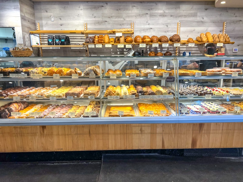 inside of bakery with large glass case of baked goods and bread on shelves on wall behind.