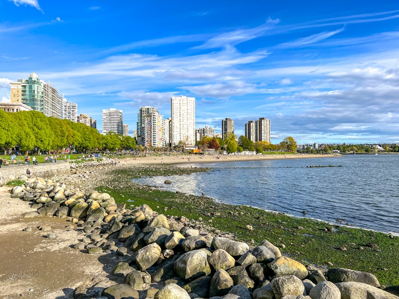 rocky shoreline with beach and tall city buildings in background with blue sky above.