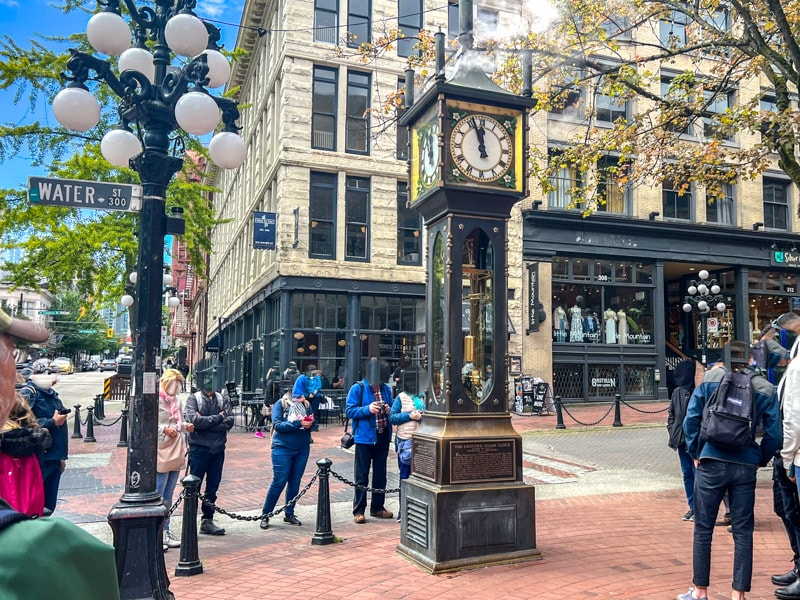 old steam clock with many people standing around it on historic street corner.