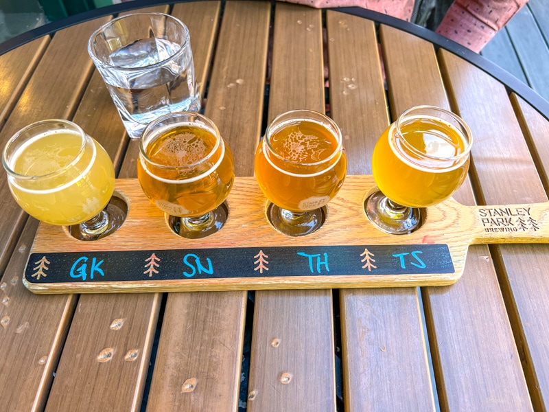 four small sampler beer in wooden paddle on table with water glass behind.