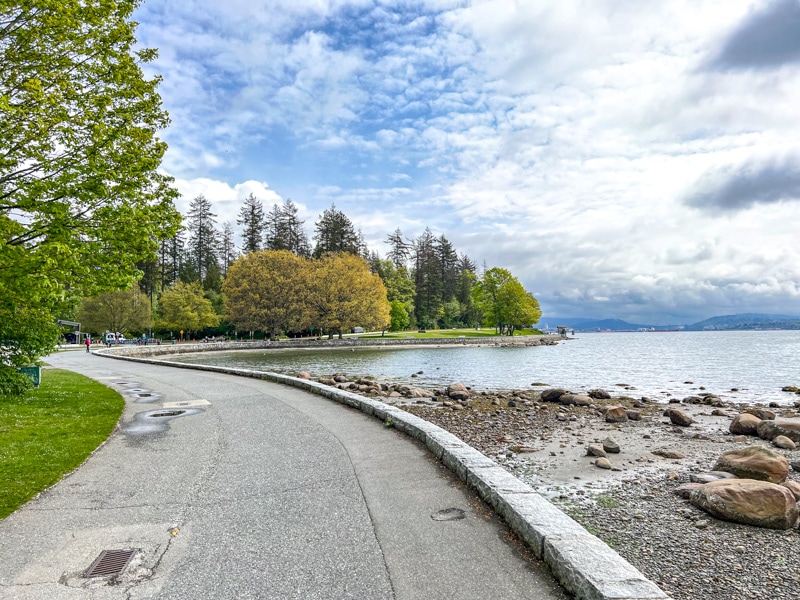 long waterfront path with rocky shoreline and forest trees along under blue cloudy sky.